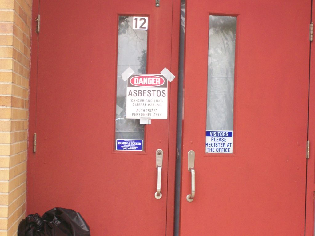 An asbestos warning sign on red school doors, representing the risk of toxic exposure in aging Louisiana public buildings.